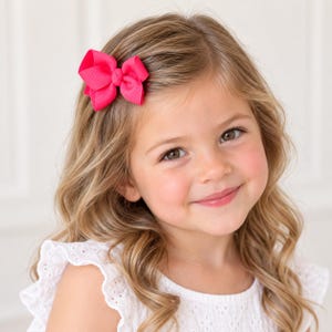 May include: A bright hot pink hair bow made of ribbed grosgrain ribbon. The bow features a classic layered design with a central knot detail, securely attached to the side of light brown wavy hair, providing a vibrant pop of color.