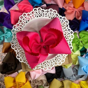 May include: A bright hot pink grosgrain ribbon hair bow with scalloped edges, centered on a white circular lace-patterned doily, surrounded by a large collection of similar hair bows in various solid colors including yellow, blue, and green.