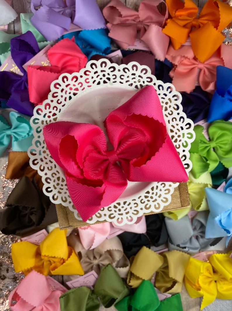May include: A bright hot pink grosgrain ribbon hair bow with scalloped edges, centered on a white circular lace-patterned doily, surrounded by a large collection of similar hair bows in various solid colors including yellow, blue, and green.