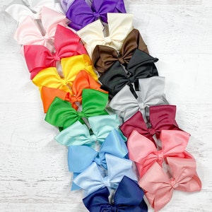May include: A collection of 18 large satin bows in various colors, including purple, white, red, yellow, orange, green, blue, and pink. The bows are arranged in a cascading pattern on a white wooden surface.