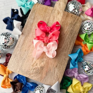 May include: A set of three pink, red, and light pink hair bows on a wooden cutting board. The bows are made of satin ribbon and are tied with a knot in the center. There are several other hair bows in various colors around the cutting board.