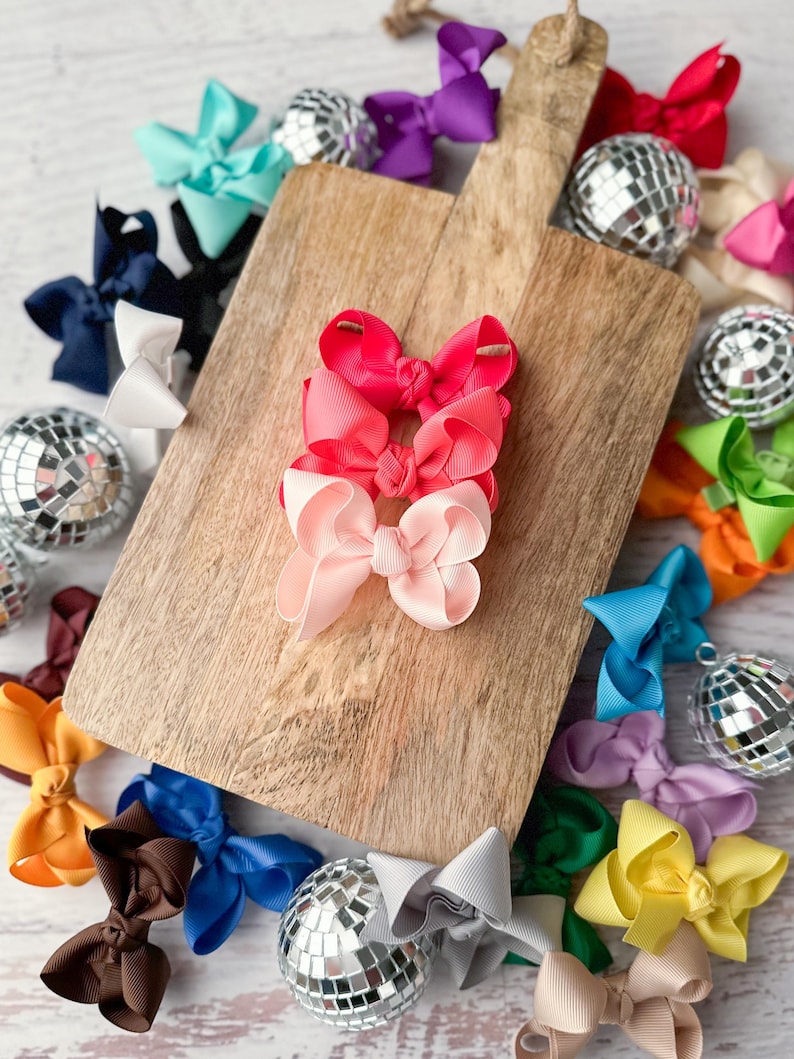 May include: A set of three pink, red, and light pink hair bows on a wooden cutting board. The bows are made of satin ribbon and are tied with a knot in the center. There are several other hair bows in various colors around the cutting board.
