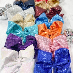 May include: A collection of large, sequined hair bows in various colors, including silver, gold, red, blue, and orange. The bows are arranged in rows, with a few disco ball ornaments scattered around them. The bows are likely for girls or women.