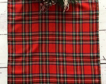 May include: A red plaid table runner with a Christmas theme. The runner features a classic tartan pattern in red, black, white, and yellow. A pine cone, evergreen sprigs, and red berries adorn the top of the runner.