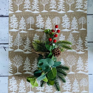 May include: A tan table runner with a repeating pattern of white winter trees. A decorative arrangement of greenery, red berries, and a pinecone sits on top. The runner is made of a textured fabric, perfect for holiday decor.