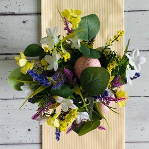 May include: A vibrant Easter display with pink speckled eggs nestled amongst white, yellow, and purple flowers and green leaves. The arrangement sits on a yellow and white striped backdrop, creating a cheerful, springtime feel.