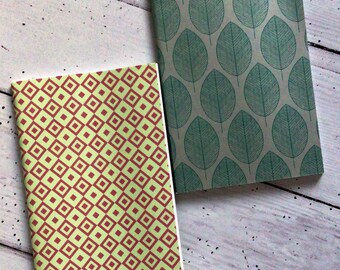 May include: Two notebooks with patterned covers. One has a light green background with a repeating pattern of red diamonds. The other has a grey background with a teal leaf pattern. The notebooks are on a light wood surface.