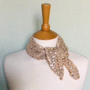May include: A gold sequined scarf tied around a mannequin's neck. The scarf is made of small, shimmering sequins and has a loose, flowing shape.