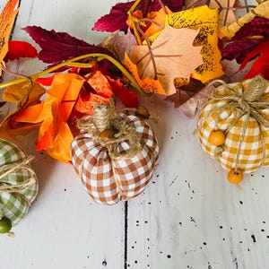 May include: Three fabric pumpkins in green, brown, and yellow gingham patterns, each topped with a jute twine stem and tied with twine featuring small wooden beads. The pumpkins are arranged in front of a garland of artificial autumn leaves.