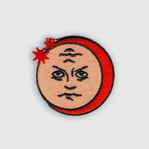 Puede incluir: Parche bordado en rojo y rosa de una cara con el ceño fruncido y una forma de media luna alrededor de la cabeza. Dos estrellas rojas están por encima de la cabeza.