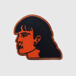 May include: An orange embroidered patch of a woman's face with black hair and stitched-up eyes and mouth.