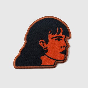Könnte beinhalten: Ein orangefarbener gestickter Aufnäher einer Person mit schwarzen Haaren, die nach rechts schaut.