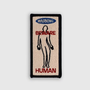 Op de afbeelding: Een beige geborduurde patch met een zwarte rand. De patch heeft een waarschuwingsbord met de tekst "WARNING" in blauw, "BEWARE" in rood en "HUMAN" in rood. Een zwart silhouet van een persoon bevindt zich in het midden van de patch.