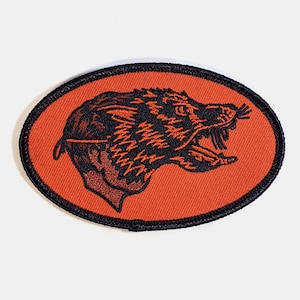 Könnte beinhalten: Ein ovales Abzeichen mit schwarzem Rand, das einen orangefarbenen Hintergrund und eine schwarze Illustration aufweist. Die Illustration zeigt den Kopf eines knurrenden Wolfes und das Profil einer Person. Das Abzeichen befindet sich auf weißem Hintergrund.