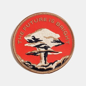 May include: A round, embroidered patch with a red background and a beige mushroom cloud. The text "THE FUTURE IS BRIGHT" is embroidered in gold around the mushroom cloud.