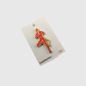 May include: A red devil-shaped pin with gold accents, featuring a stylized figure in a dynamic pose. The pin is attached to a light-colored card with the word "PIN" and the brand name "badaboom".