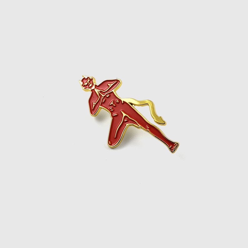 May include: A red enamel pin in the shape of a stylized figure with a devilish appearance. The pin has a gold-colored outline and details. The figure is in a running pose with arms raised.