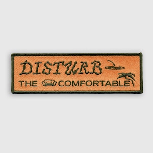 Może przedstawiać: Naszywka haftowana w kolorze brązowym i zielonym z tekstem "Disturb The Comfortable" i małym wzorem sofy i palmy.