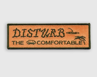 Puede incluir: Un parche bordado marrón y verde con el texto "Disturb The Comfortable" y un pequeño gráfico de un sofá y una palmera.