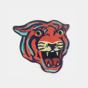 May include: An embroidered iron-on patch featuring a red tiger head with a blue outline and yellow teeth. The tiger is roaring with its mouth open.