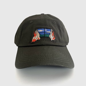 May include: A dark olive green baseball cap with a curved brim. The cap features an embroidered patch of a window scene with blue sky, green hills, and flames on either side. A casual accessory.