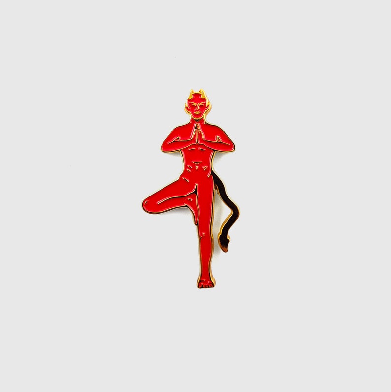 May include: A red enamel pin featuring a stylized devil figure in a yoga pose. The pin has a gold outline, a black tail, and a red body with yellow horns. The devil is standing on one leg with hands together.