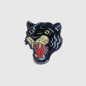May include: Embroidered patch featuring a roaring black panther head. The patch has a detailed design with yellow, red, purple, and teal accents. The panther's mouth is open, revealing sharp teeth and a red interior.