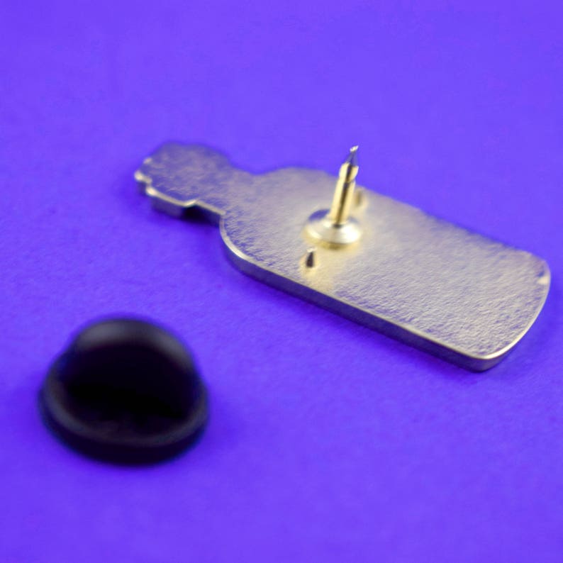 May include: The back of a gold-coloured enamel pin shaped like a bottle. The pin has a black rubber clutch on the back.
