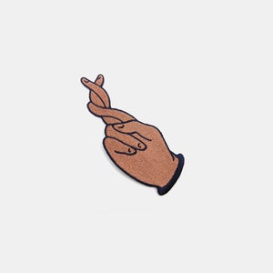 May include: Embroidered patch featuring a hand with fingers crossed in a gesture of luck. The patch has a brown hand with black outlines and a navy blue cuff. The design is on a white background.
