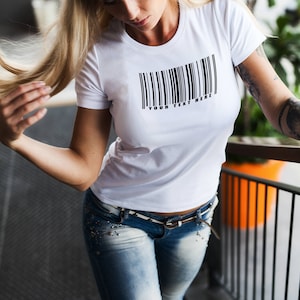 以下が含まれることがあります： 黒いバーコードのグラフィックと「YOUR TEXT HERE」というテキストが前面に印刷された白いTシャツ。長いブロンドの髪と青いジーンズを履いた人が着用しています。