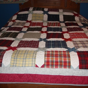 May include: A patchwork quilt with a red, white, and blue plaid pattern. The quilt is made of different fabrics and is quilted with a white thread. The quilt is on a bed with a wooden headboard.