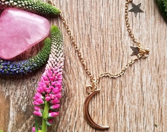Könnte beinhalten: Eine goldene Halskette mit einem Halbmond-Anhänger und zwei Sternen-Anhängern. Die Halskette liegt auf einer Holzoberfläche mit rosa und grünen Blumen.