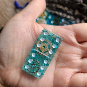 Op de afbeelding: Een turquoise blauwe domino met witte stippen en gouden glitter. De domino heeft een gouden tandwielontwerp.