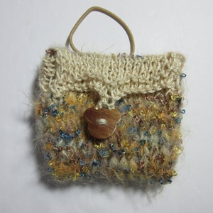 May include: A small, handmade knitted bag with a cream-colored top and a multi-colored body. The body of the bag features a mix of blue, gold, and brown yarns. A wooden flower-shaped button secures the flap, and a brown elastic loop serves as a handle.