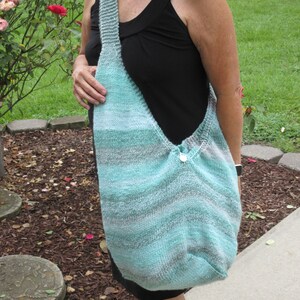 May include: A large, knitted shoulder bag in shades of teal, gray, and white. The bag has a wide strap and a button closure. The knitted texture is visible, and the bag appears to be handmade. The bag is worn over the shoulder.