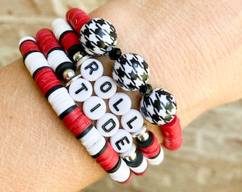 May include: A set of four bracelets featuring red, white and black beads. One bracelet has the word "ROLL TIDE" spelled out in white beads. Another bracelet has black and white houndstooth patterned beads. The bracelets are stacked on a person's wrist.