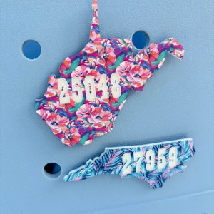 May include: Two state-shaped shoe charms with floral and tropical patterns. The West Virginia charm has a pink, blue, and green floral pattern with the number "25048" in white. The North Carolina charm has a blue, pink, and green tropical leaf pattern with the number "27959" in white. The charms are on a light blue background.