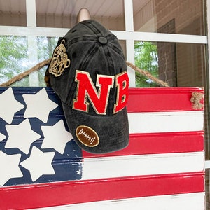 May include: A black baseball cap with the letters "NB" in red and gold trim, and a football patch. The cap is hanging on a hook above a wooden American flag with red and white stripes and white stars.