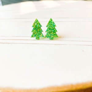 May include: Two green glitter Christmas tree stud earrings. The earrings are shaped like stylized evergreen trees with a textured, sparkly appearance. The earrings are set against a white background.