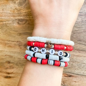 May include: A stack of bracelets with red, white, and black beads. The top bracelet has a silver star charm and the bottom bracelet has the word "OHIO" spelled out in white letters.