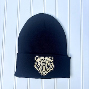 May include: A black beanie hat with a gold embroidered bear patch on the front. The hat is made of a soft, knitted material and has a folded brim. The bear patch features a detailed design of a roaring bear.