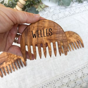 May include: Wooden comb with the name "WELLES" in black lettering. The comb is made of a light brown wood with a natural grain pattern. The comb has a curved top and several teeth. Additional combs are visible in the background.