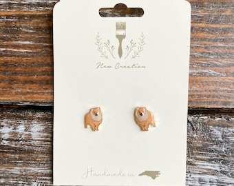 May include: A pair of gold stud earrings featuring a small, cartoon-style illustration of a brown and white dog. The earrings are displayed on a white card with the text "New Creation" and "Handmade in NC".