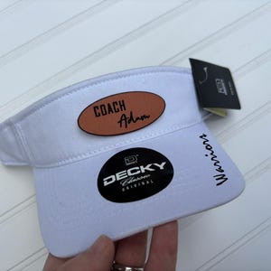 May include: White visor with the text "COACH Adam" on a brown oval patch. The visor also has the text "Warriors" and a "DECKY Classic Original" logo. A black tag is attached.