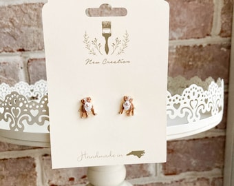 May include: A pair of gold stud earrings featuring a brown and white dog design. The earrings are displayed on a white stand with a lace trim. The card behind the earrings reads "New Creation" and "Handmade in NC".