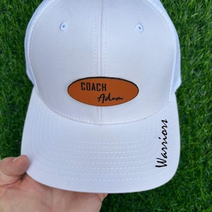 May include: White trucker hat with a curved brim and mesh back. Features a brown oval patch with the text "COACH Adam" and the word "Warriors" printed on the side in black. The hat is held against a green grass background.