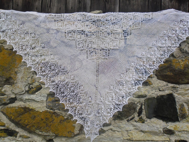 May include: A white crocheted shawl with a delicate lace pattern. The shawl is draped over a stone wall.