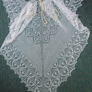May include: A white crocheted shawl with a delicate lace pattern. The shawl is draped over a green surface and has a strand of pearls resting on top.