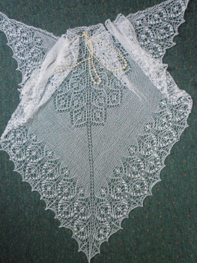 May include: A white crocheted shawl with a delicate lace pattern. The shawl is draped over a green surface and has a strand of pearls resting on top.