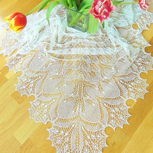 Puede incluir: Un chal de ganchillo blanco con un delicado patrón de encaje, colocado sobre una superficie de madera con un ramo de tulipanes en un jarrón de vidrio.